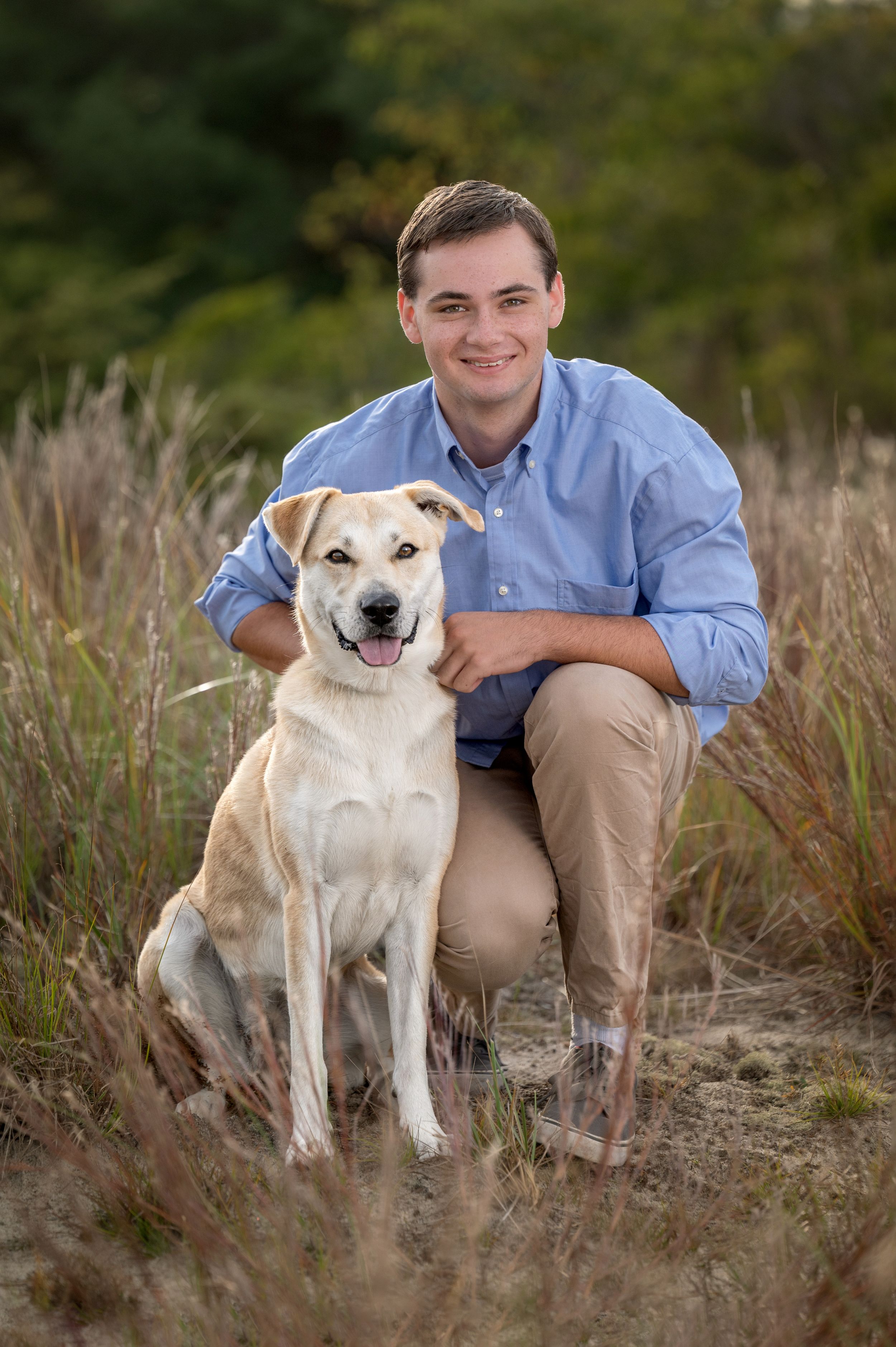A person in blue shirt poses with a happy cream-colored Labrador mix dog in a grassy outdoor setting.