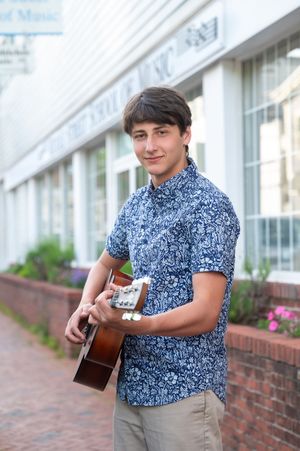 A musician in a blue floral print shirt holds an acoustic guitar while standing outside a brick building.