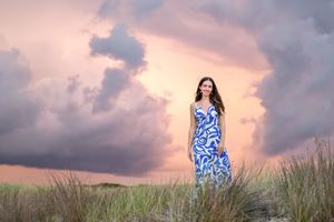 A person in a blue and white patterned dress stands among beach grass at sunset with dramatic pink clouds overhead.