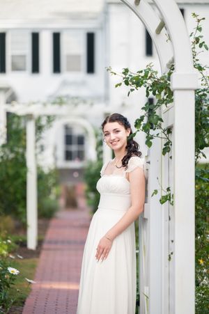 A bride in a white dress poses along a white garden arbor with climbing vines on a brick pathway.