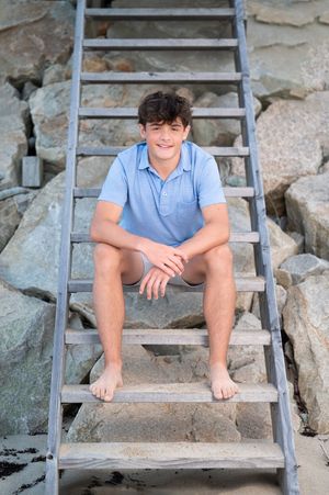 A person wearing a light blue polo shirt sits barefoot on a wooden ladder against rocky coastal terrain.