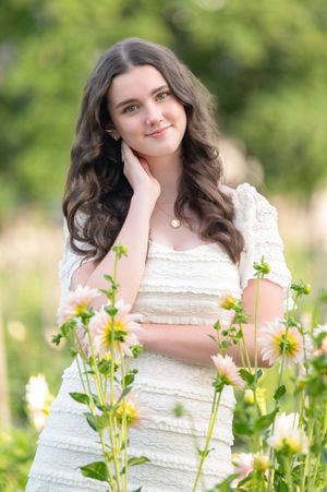 A person in a white lace dress poses among blooming yellow dahlias in a sunny garden setting.
