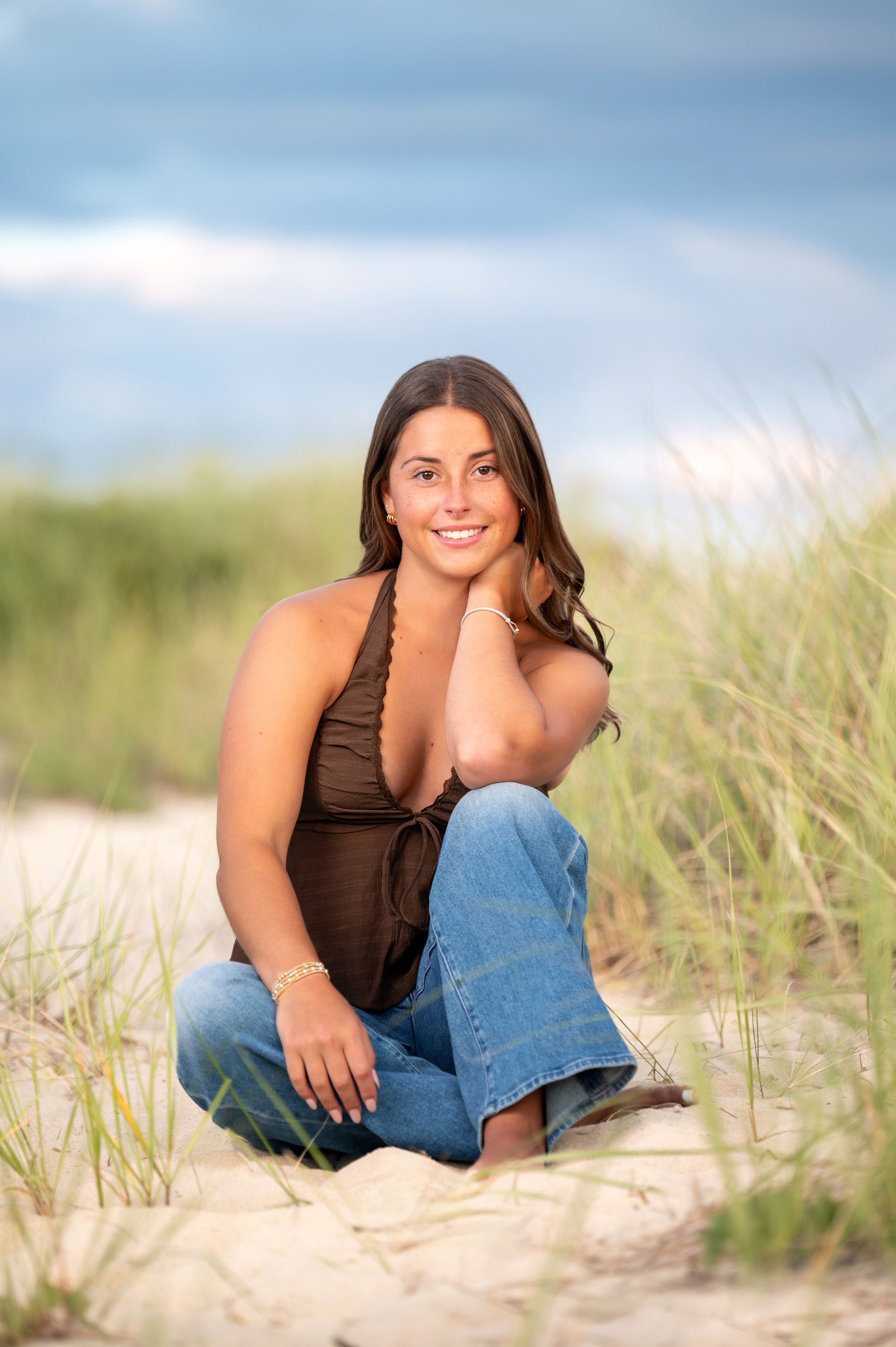 A woman in a brown tank top and jeans poses casually on a sandy beach with dune grass in the background.