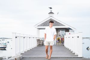 A white coastal boardwalk pier with boats in the harbor and a small building with a weathervane at the end.