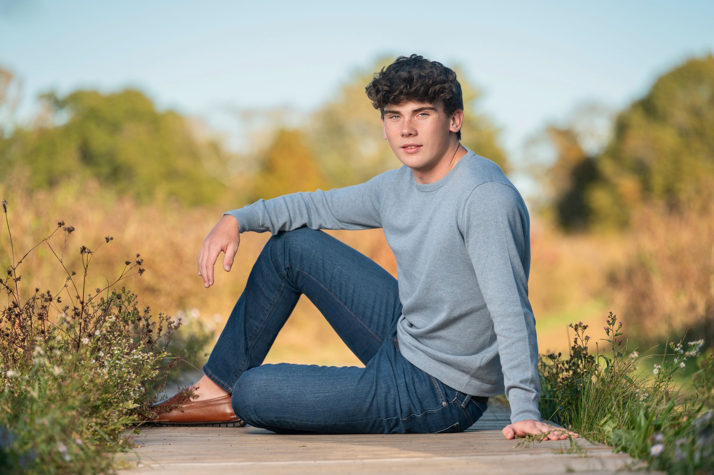 Person in gray sweater and jeans sits casually on path surrounded by autumn wildflowers and foliage.