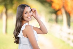 Woman in white sleeveless top smiling outdoors during golden hour with autumn colors in background.