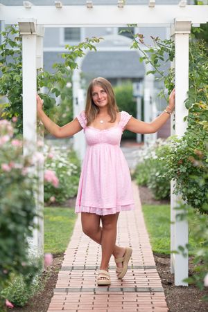 Pink sundress and platform sandals on a brick path under a white garden arbor surrounded by blooming roses and greenery.