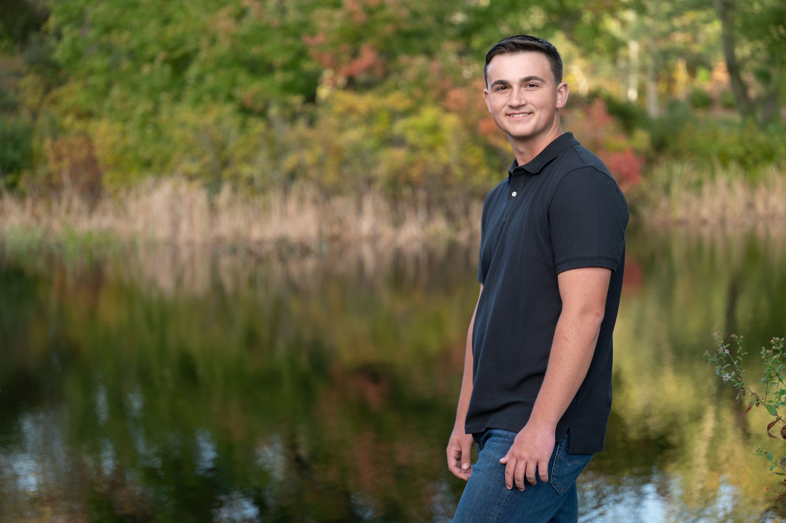 A person in a dark shirt poses outdoors near a serene lake surrounded by autumn foliage.
