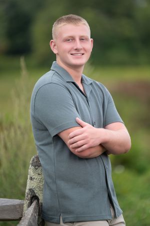 A person in a gray polo shirt poses outdoors with arms crossed, smiling against a natural green background.