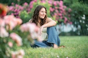 A person in a blue dress sits on grass with pink flowers blooming in the background during a sunny day.