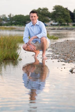 Person in blue shirt crouches near calm water's edge with reflection on rocky beach at sunset.