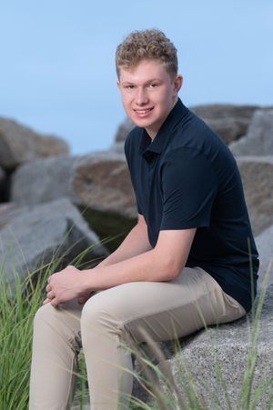 A person in a black polo shirt and khaki pants sits casually on rocky terrain with grass in the background.
