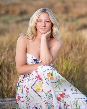 A person in a white floral sundress sits gracefully in a sunlit field of tall grass and wildflowers.