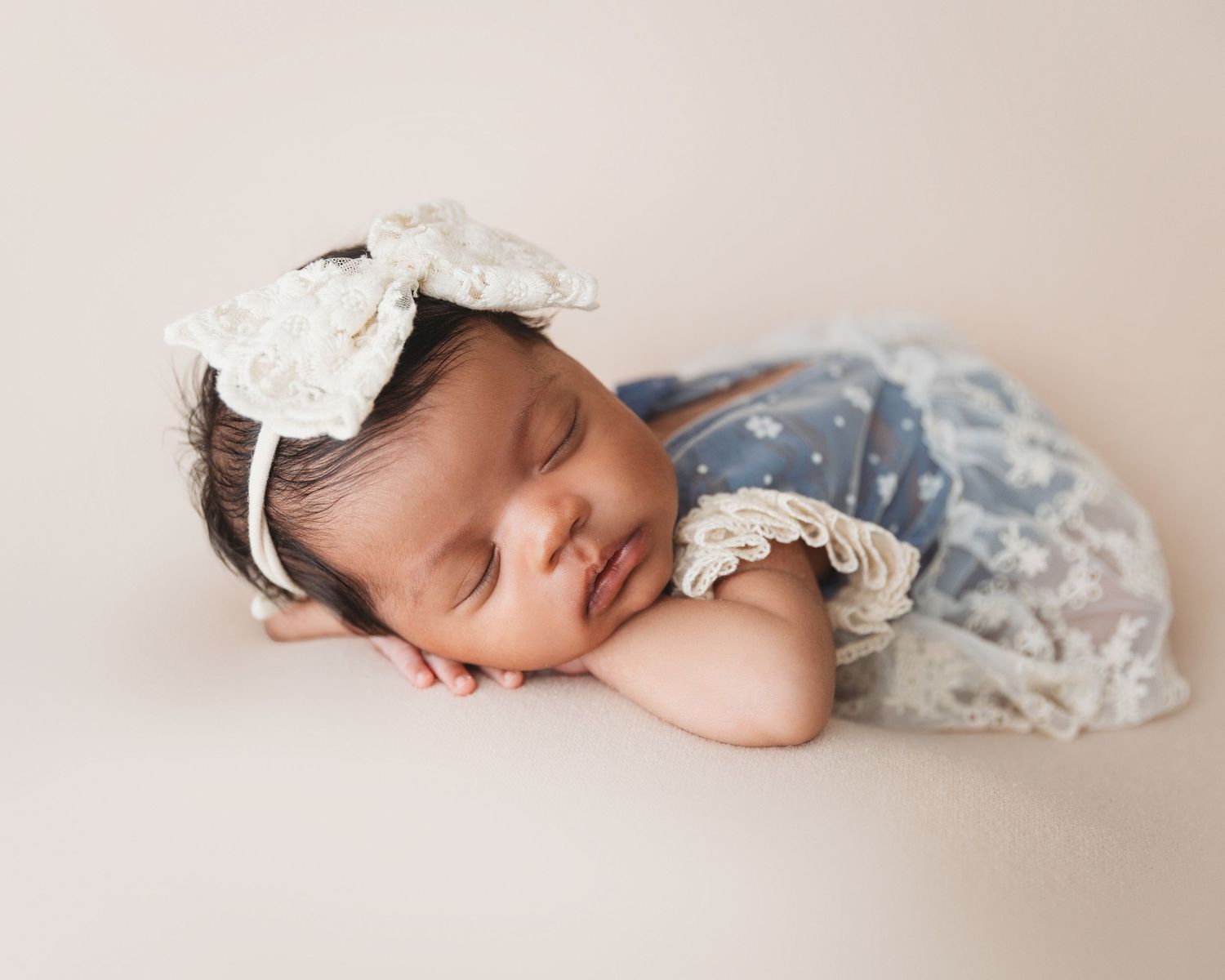 Sleeping newborn wrapped in white with delicate lace details on beige backdrop.