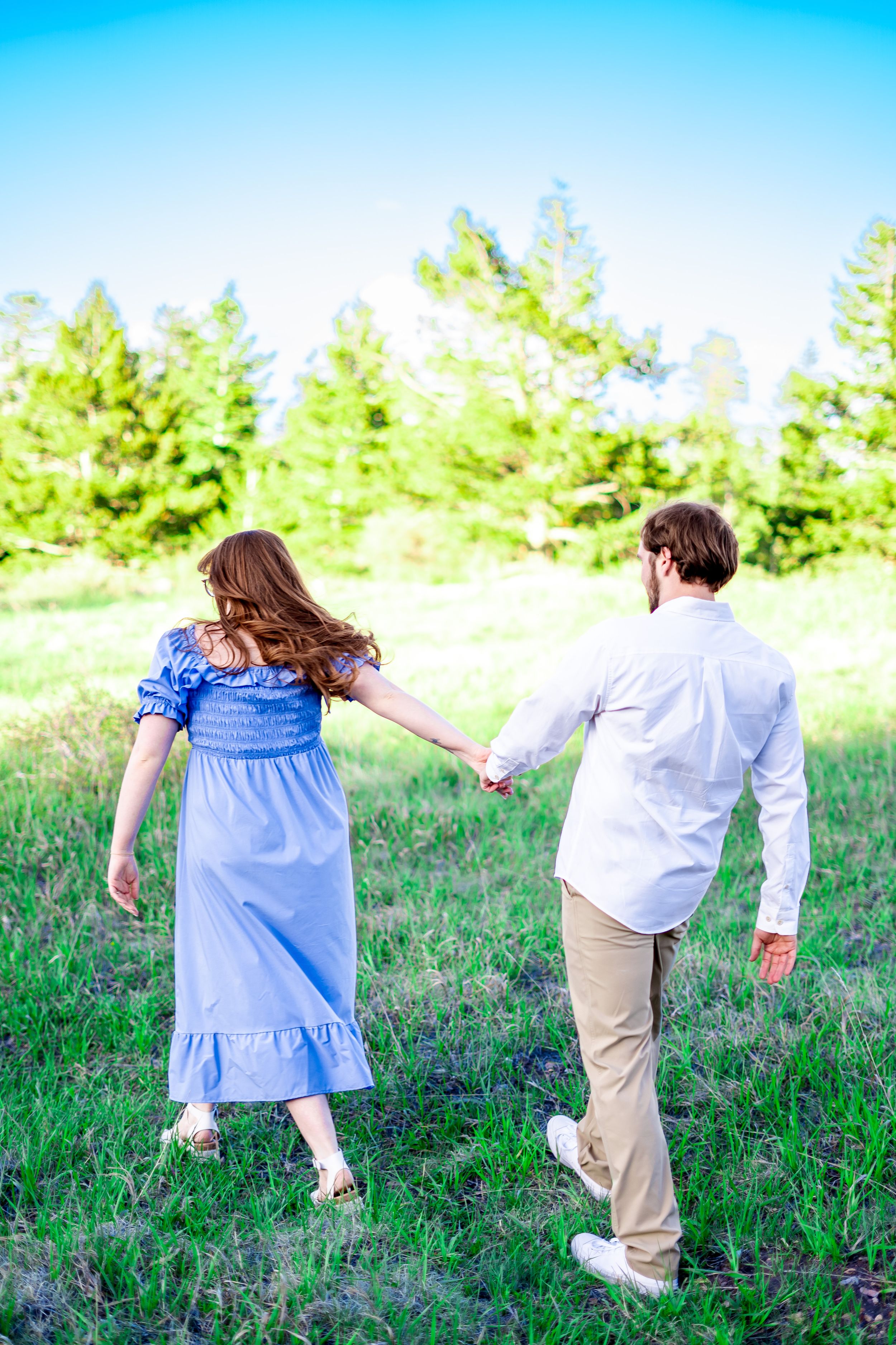 Couple holding hands while walking through a grassy meadow with evergreen trees in the background.