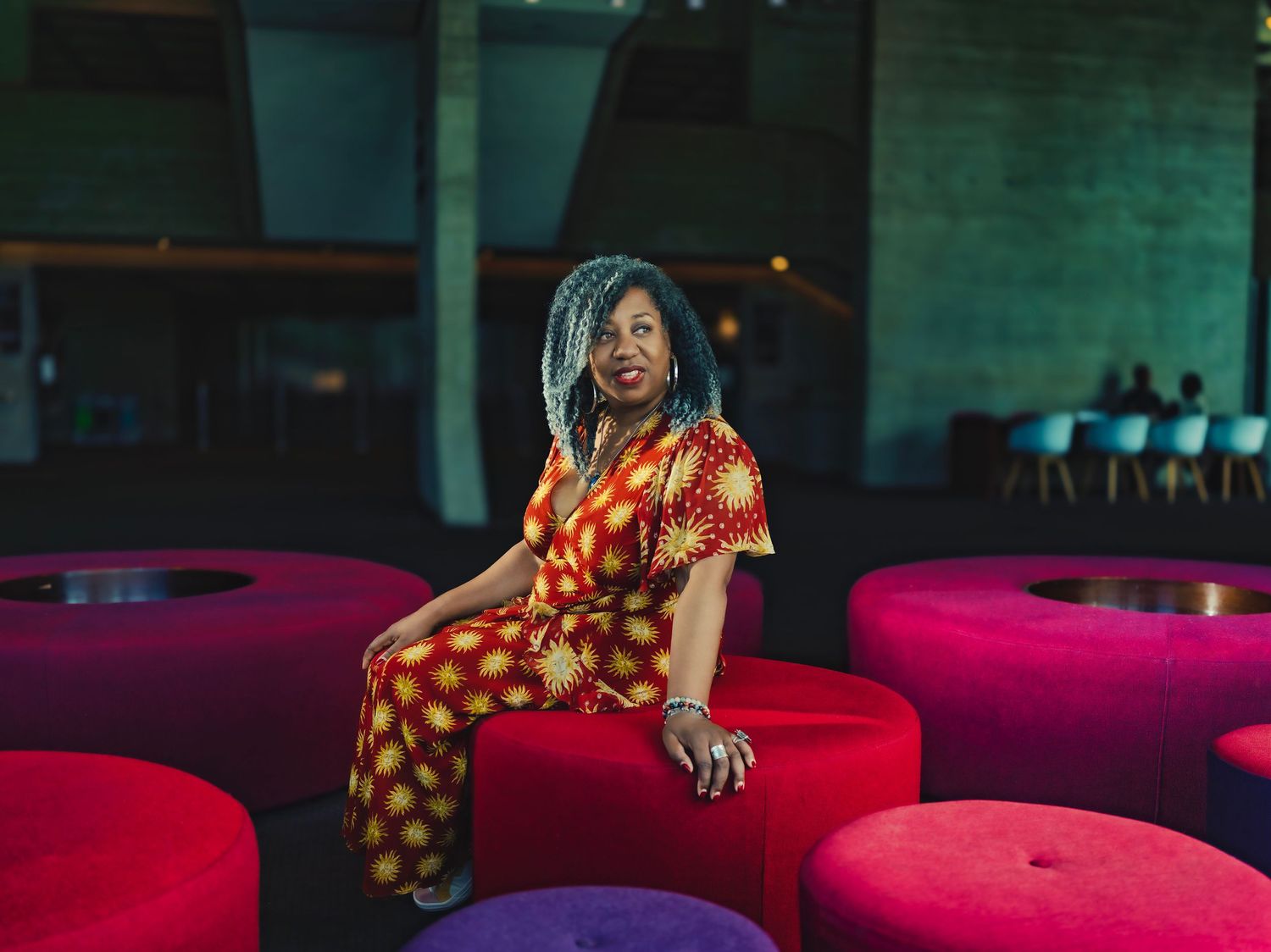 Woman in colorful patterned jumpsuit sitting on vibrant red ottoman seating.
