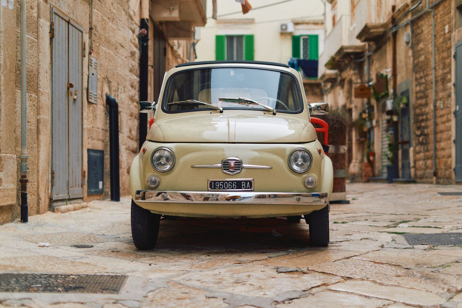 A beige vintage Fiat 500 photographed from the front on a narrow cobblestone street.