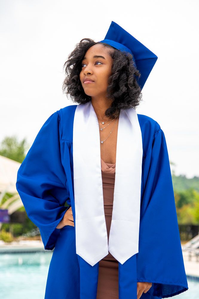 Graduate in royal blue cap and gown with white stole poses against bright outdoor background.