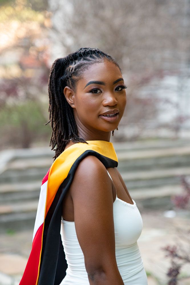 Graduate in white dress with academic regalia poses outdoors in natural lighting.