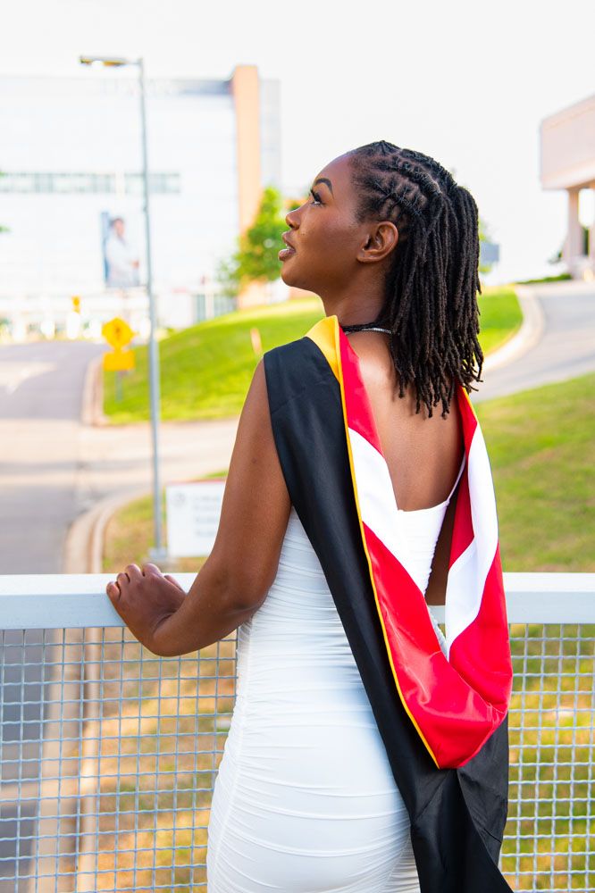 Graduate in white dress and academic stole poses against outdoor background.