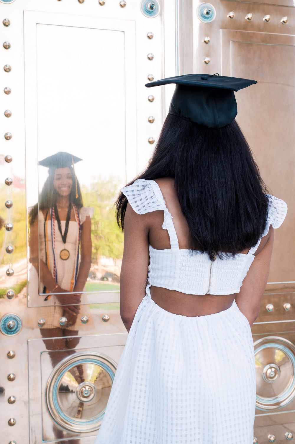 Graduate poses in a white dress against an industrial metal wall with rivets and circular details.