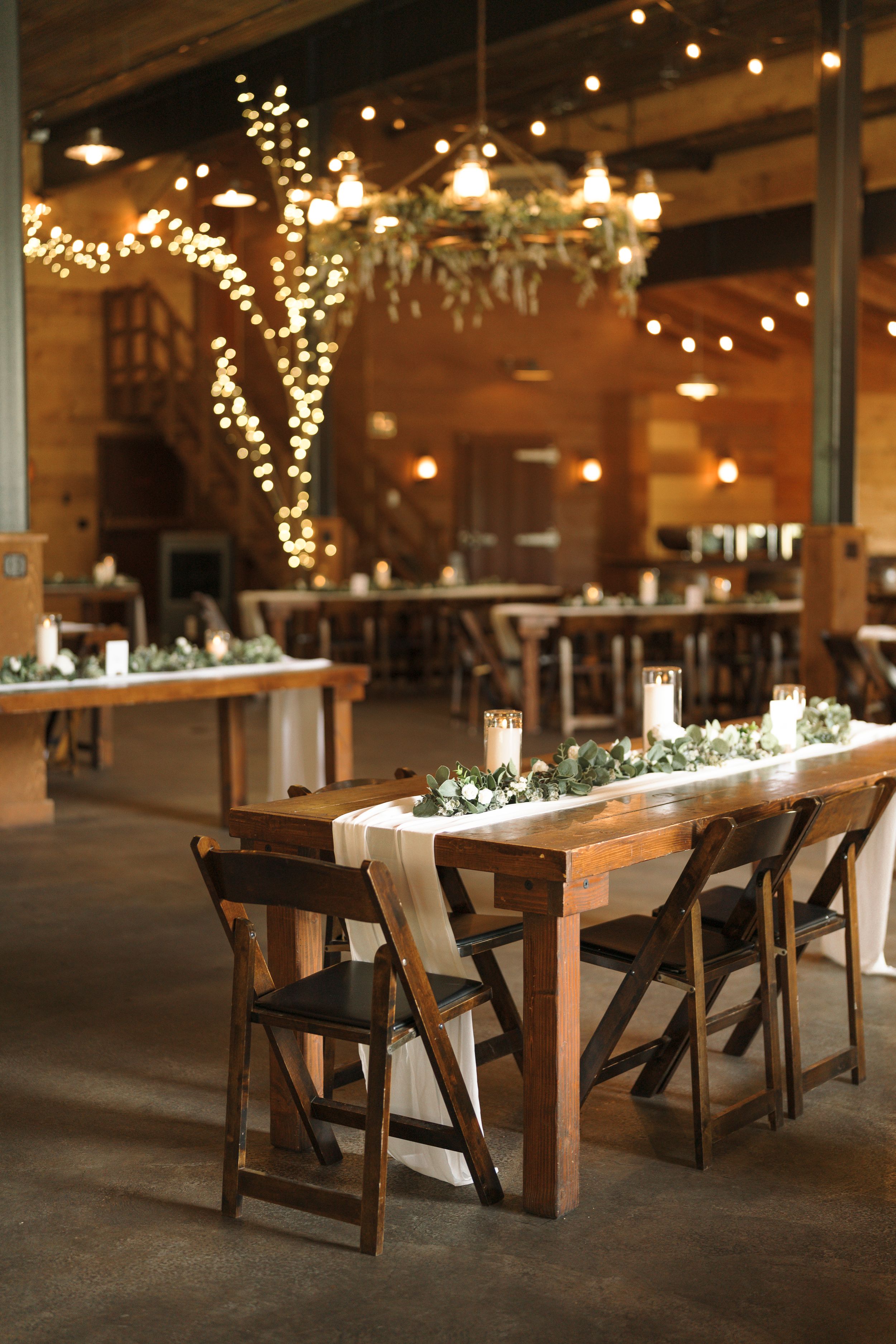 Rustic wedding reception venue with wooden tables, chairs and twinkling string lights creating a warm ambiance.