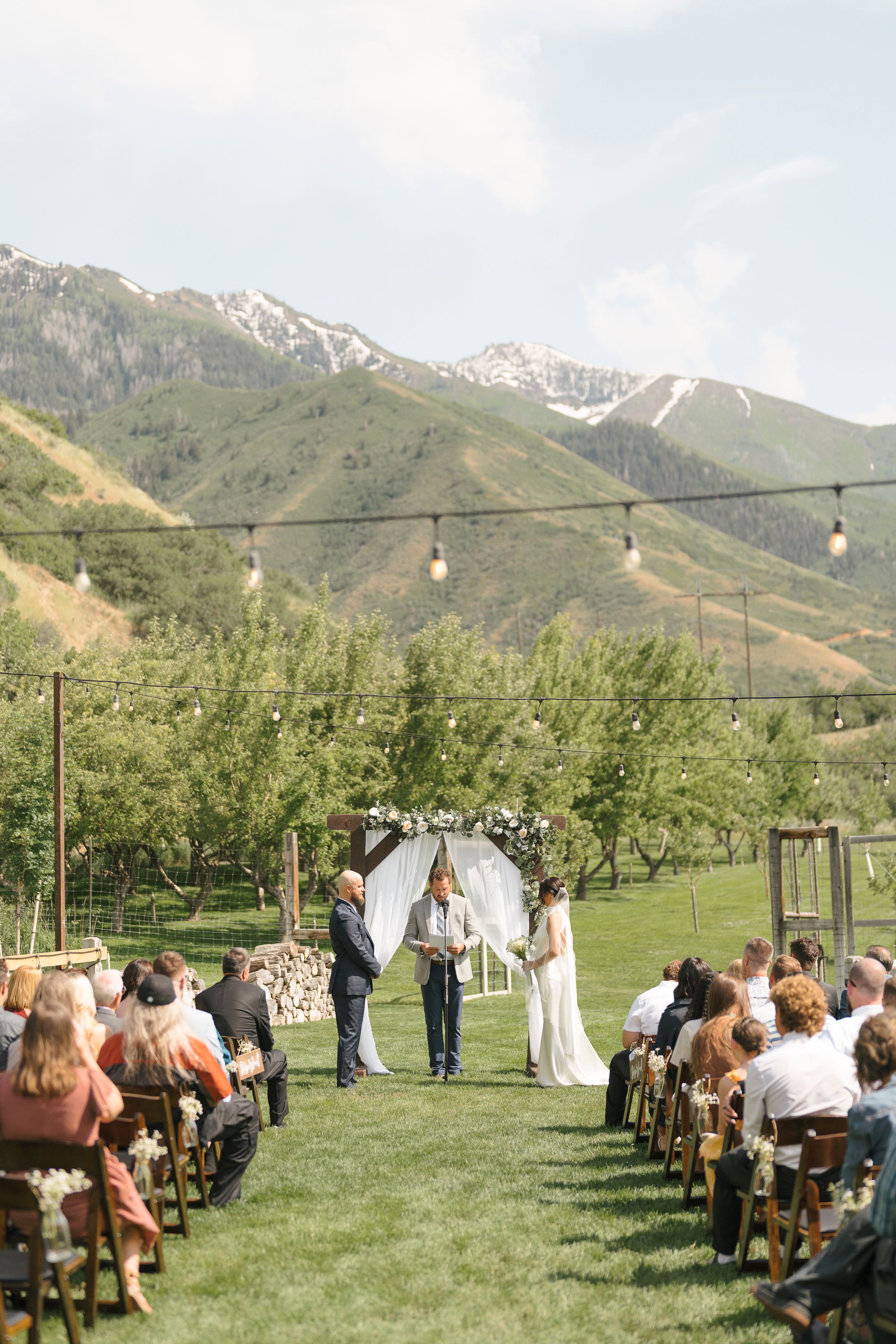 Outdoor mountain wedding ceremony with guests seated on wooden chairs surrounded by scenic mountain views.