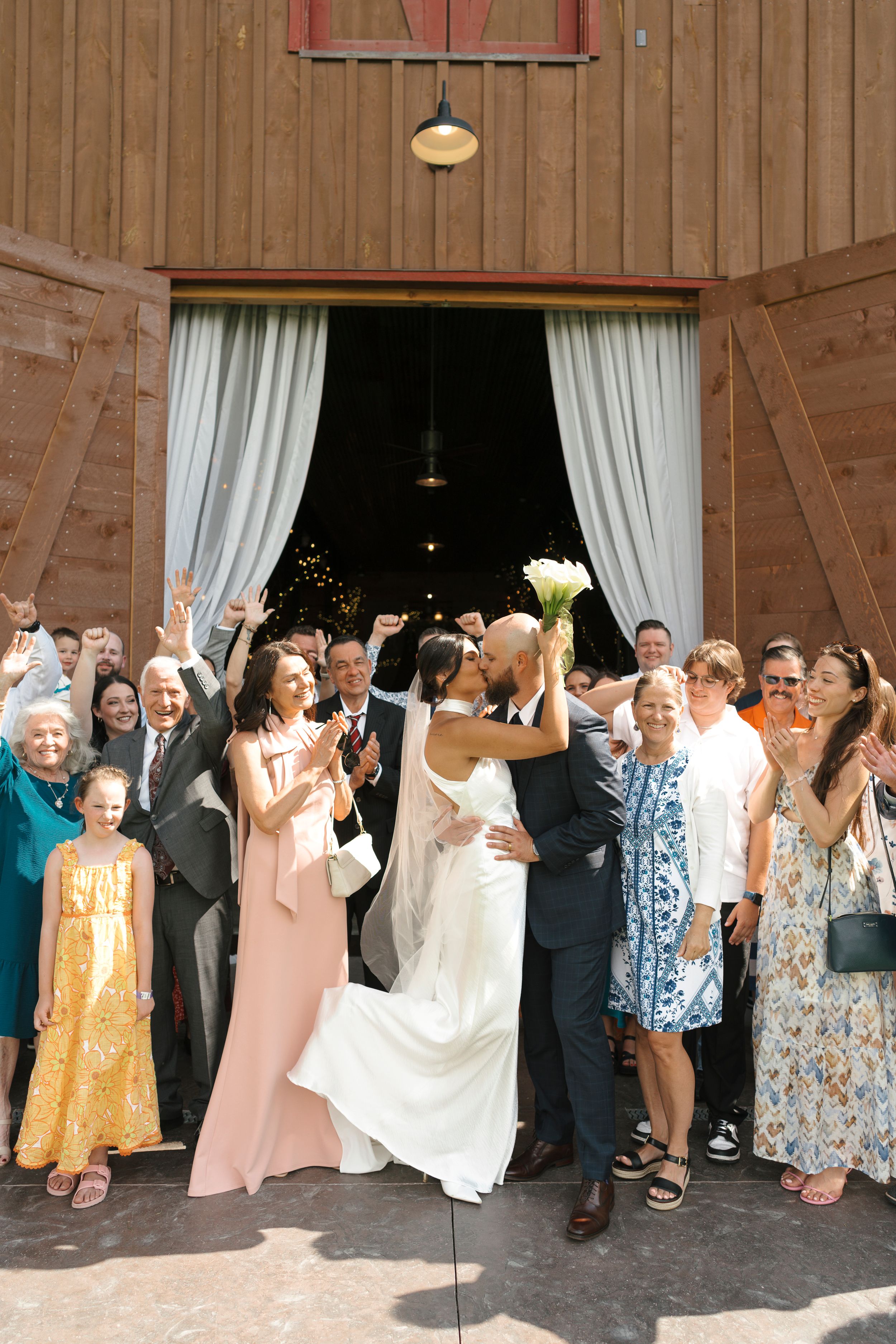 A joyful group wedding celebration with guests and the wedding party raising their hands in front of a rustic wooden barn door.