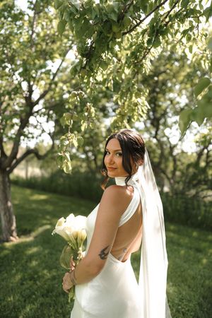 A bride in a white wedding dress poses in soft sunlight under green trees during a summer photoshoot.