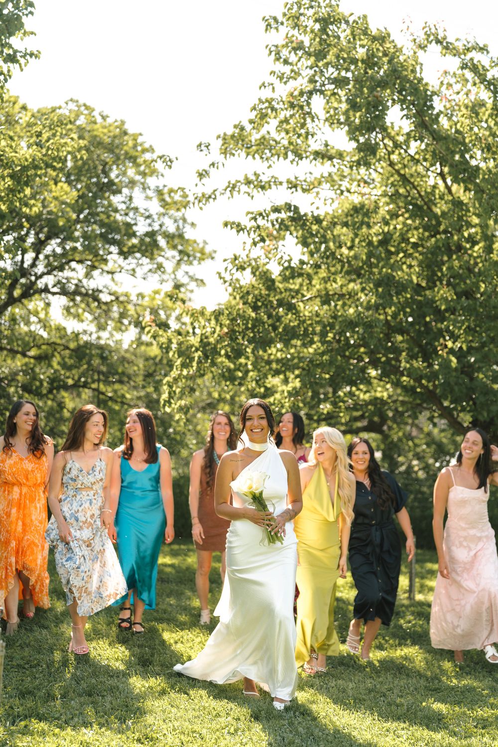A line of women in colorful dresses twirl and dance joyfully in a sunlit orchard during golden hour.