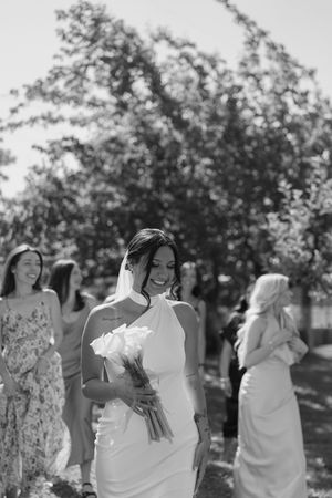 A group of joyful bridesmaids in white dresses dance and pose together outdoors on a sunny day in a garden setting.