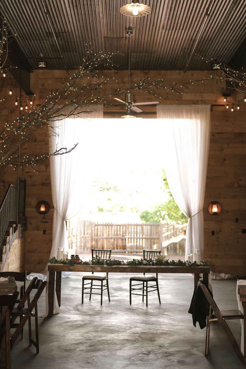 Rustic barn venue with wooden walls, string lights, and white curtains framing a bright window and two chairs at a table.