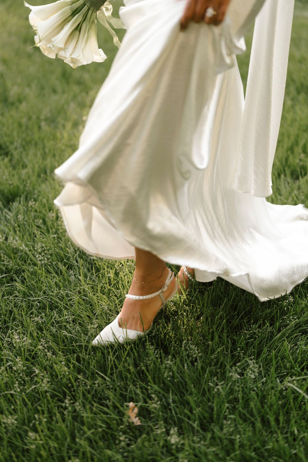 A white flowing wedding dress hemline moves above grass as bare feet with an anklet are visible underneath.