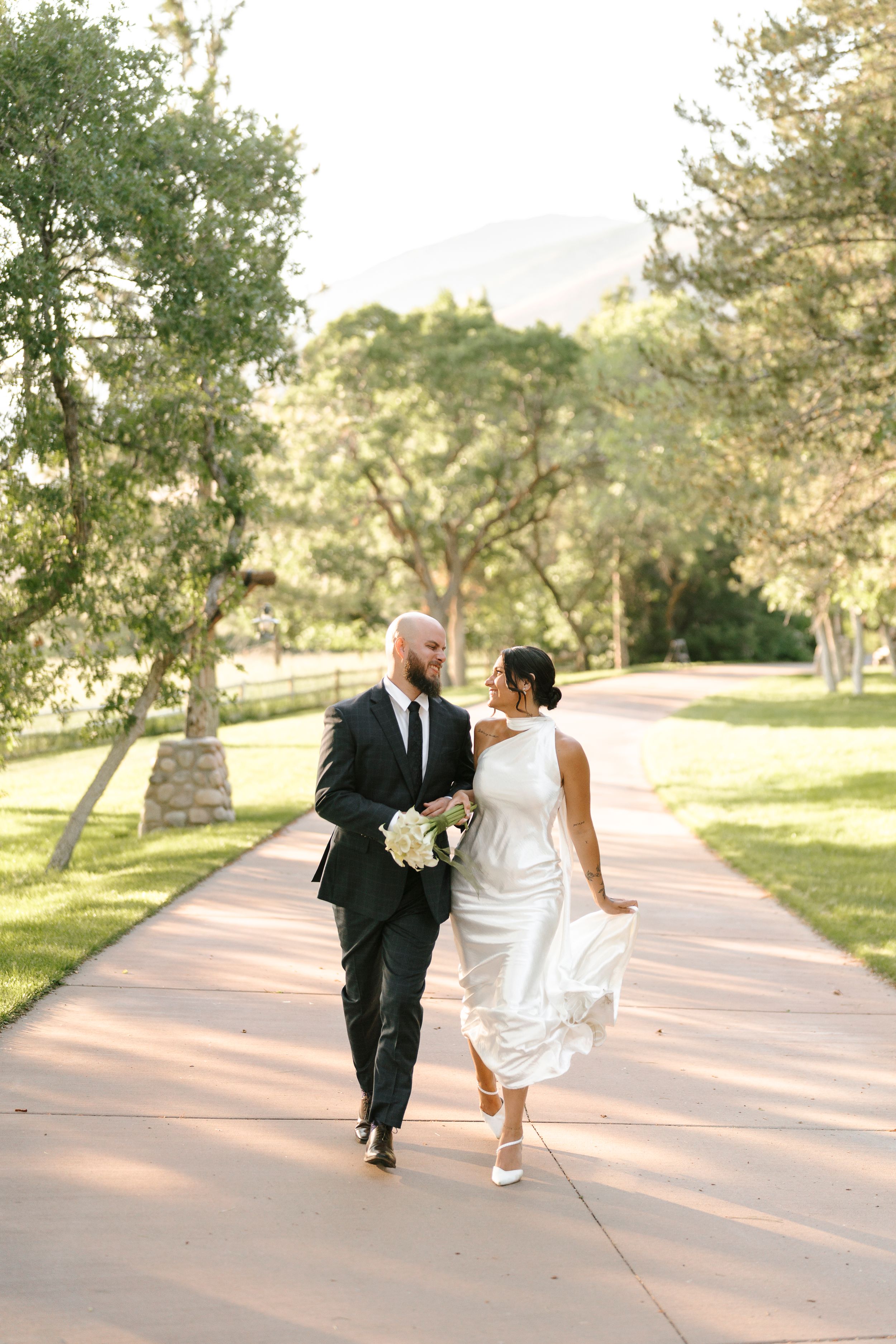 Wedding couple playfully running and walking together on sunlit pathway surrounded by green trees.
