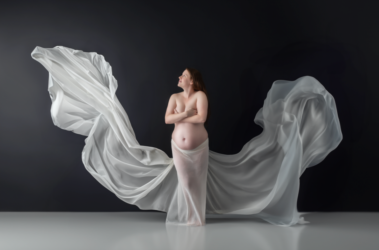 Pregnant woman draped in flowing white fabric poses gracefully against dark background in artistic maternity portrait. Embracing Life’s Beau