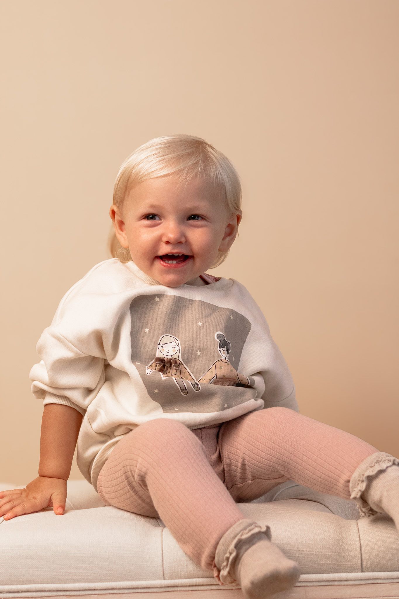 A smiling toddler wearing a cream sweatshirt with graphic print and pink leggings sits on white furniture.