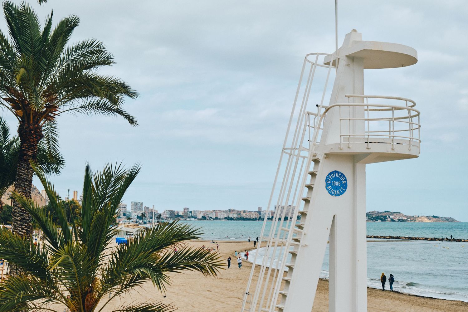 White lifeguard tower stands against a cloudy sky with palm trees and beach views in the background.