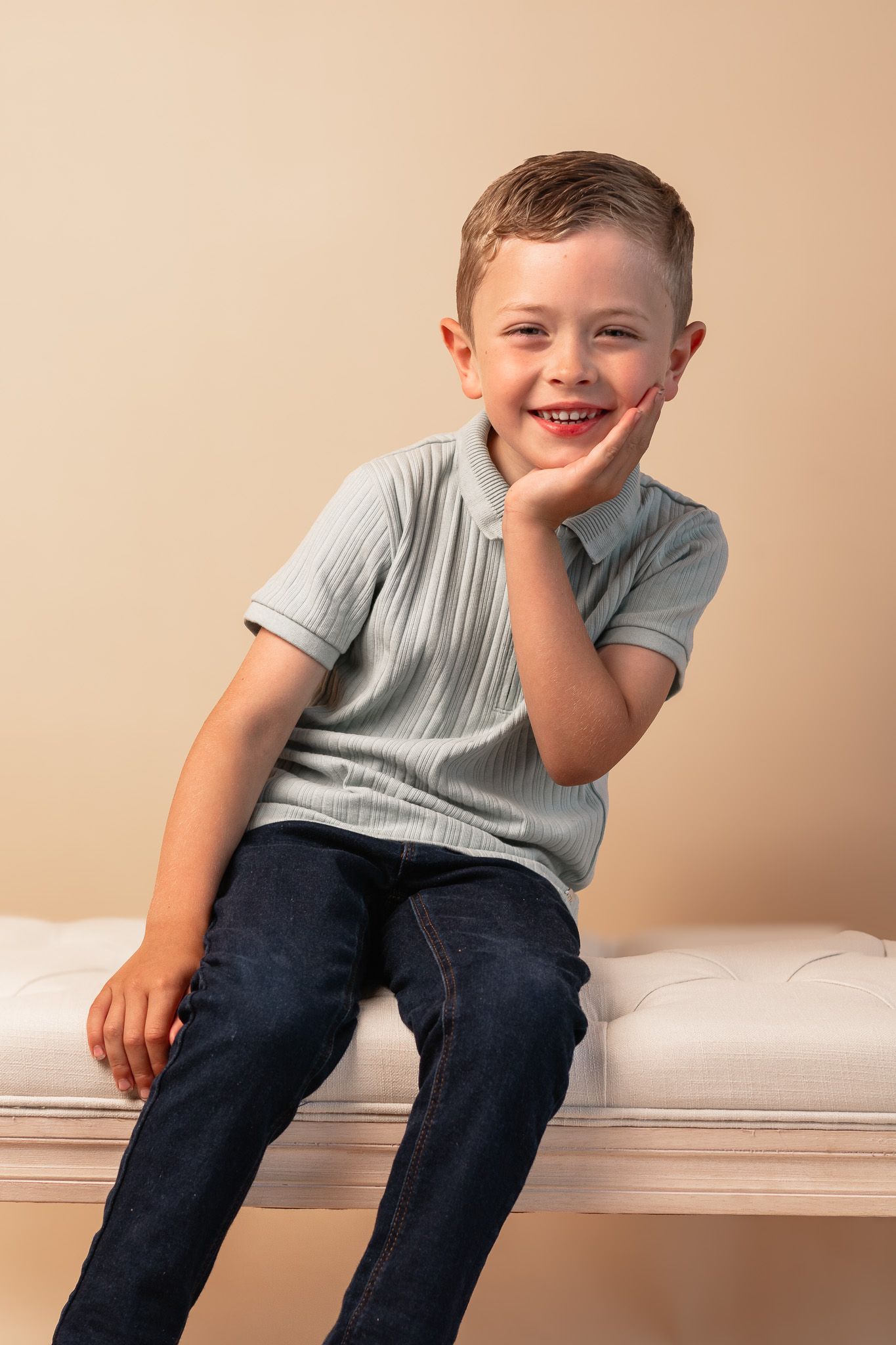 A joyful portrait of a child wearing a grey polo shirt and jeans while sitting on a white bench.