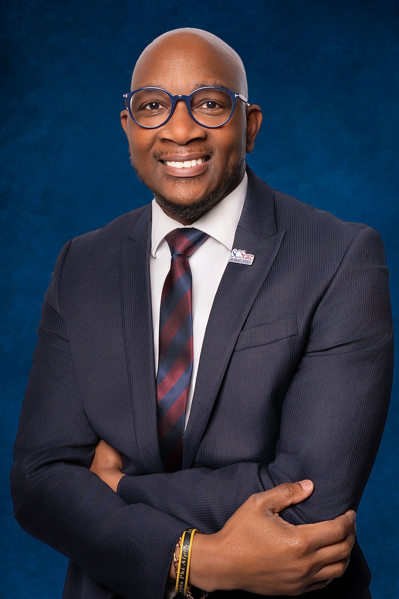 Smiling bald man in blue glasses, gray suit, and striped tie poses with arms crossed against a deep blue backdrop.