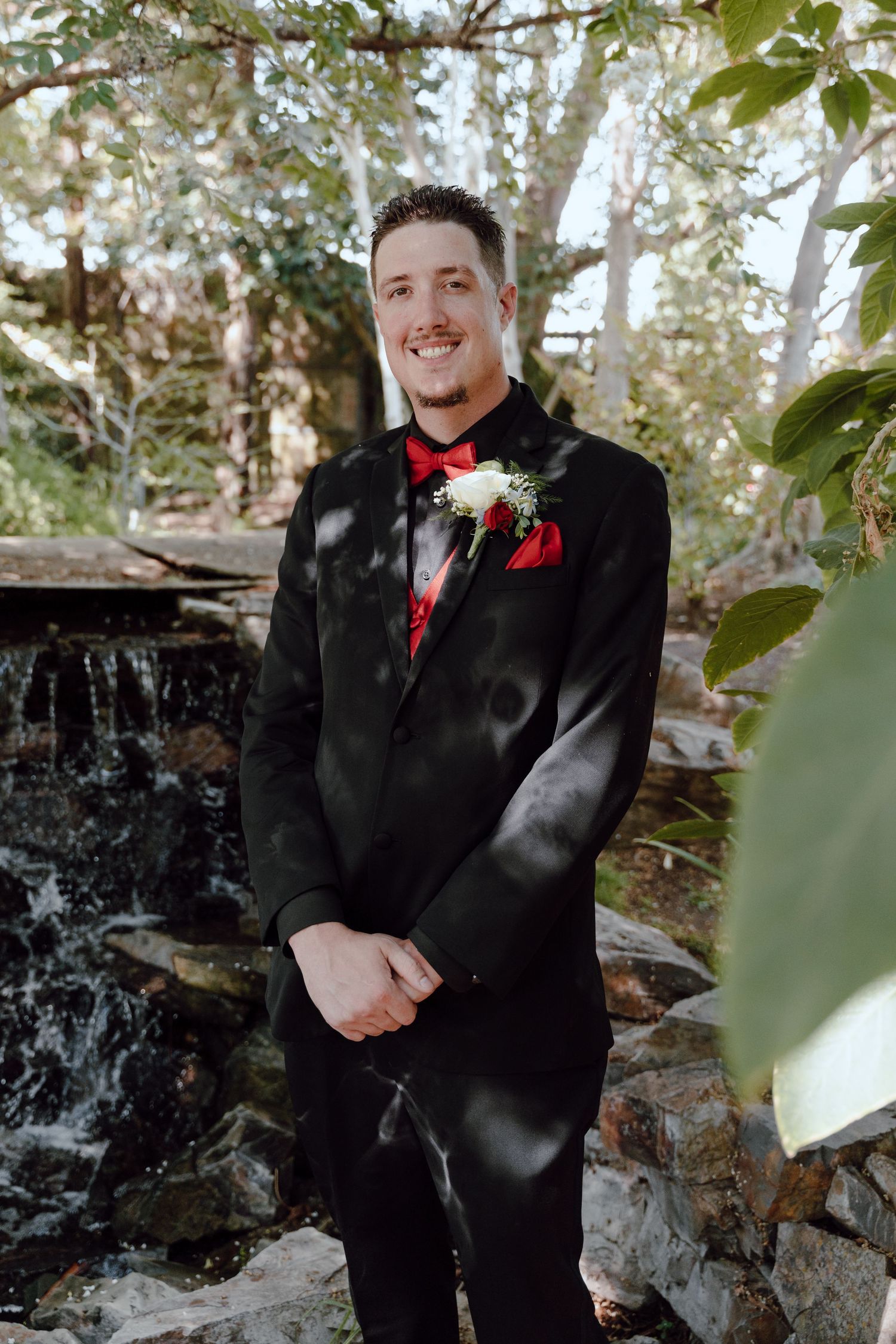 Groom in black tuxedo with red bow tie poses by garden waterfall feature.