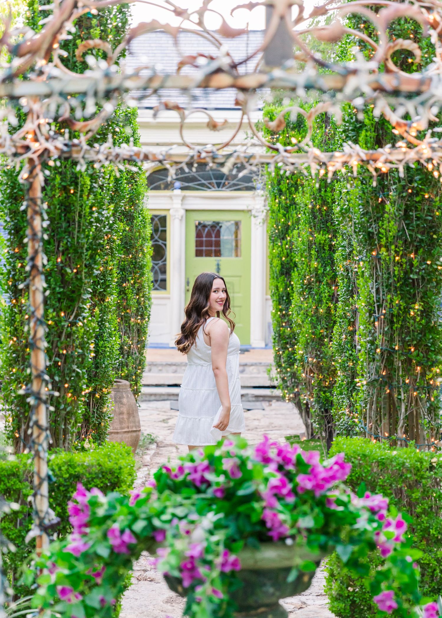 girl wearing a lacy white sundress looking over her shoulder as she walks through a hedge garden with twinkly lights