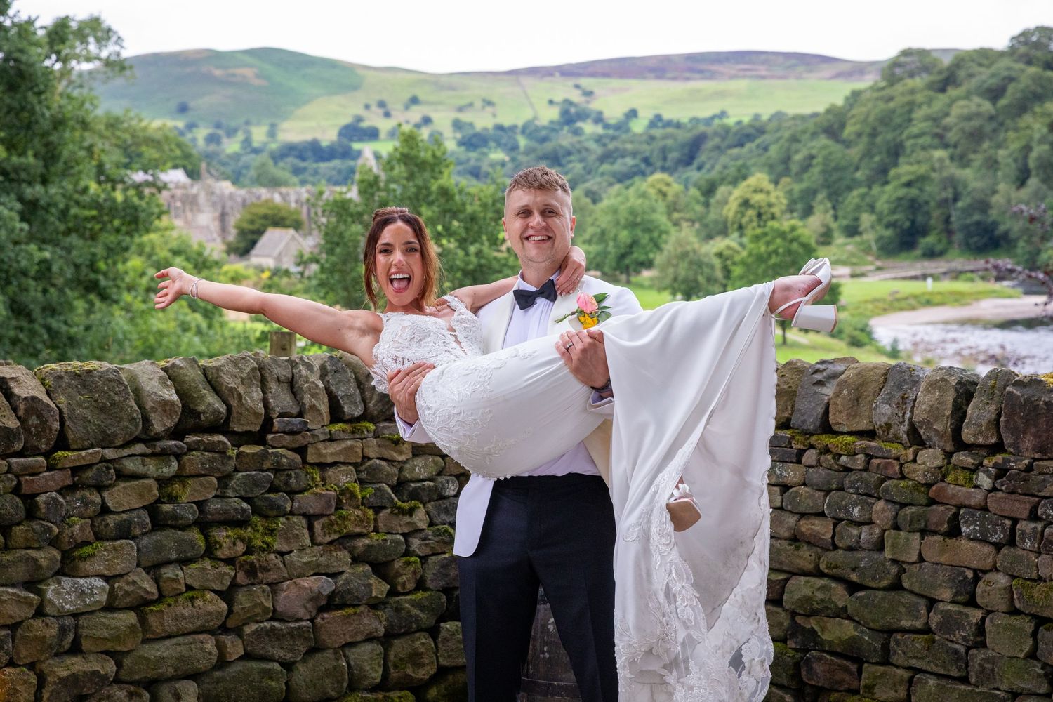 Groom carries laughing bride in white dress over stone wall with scenic valley behind.