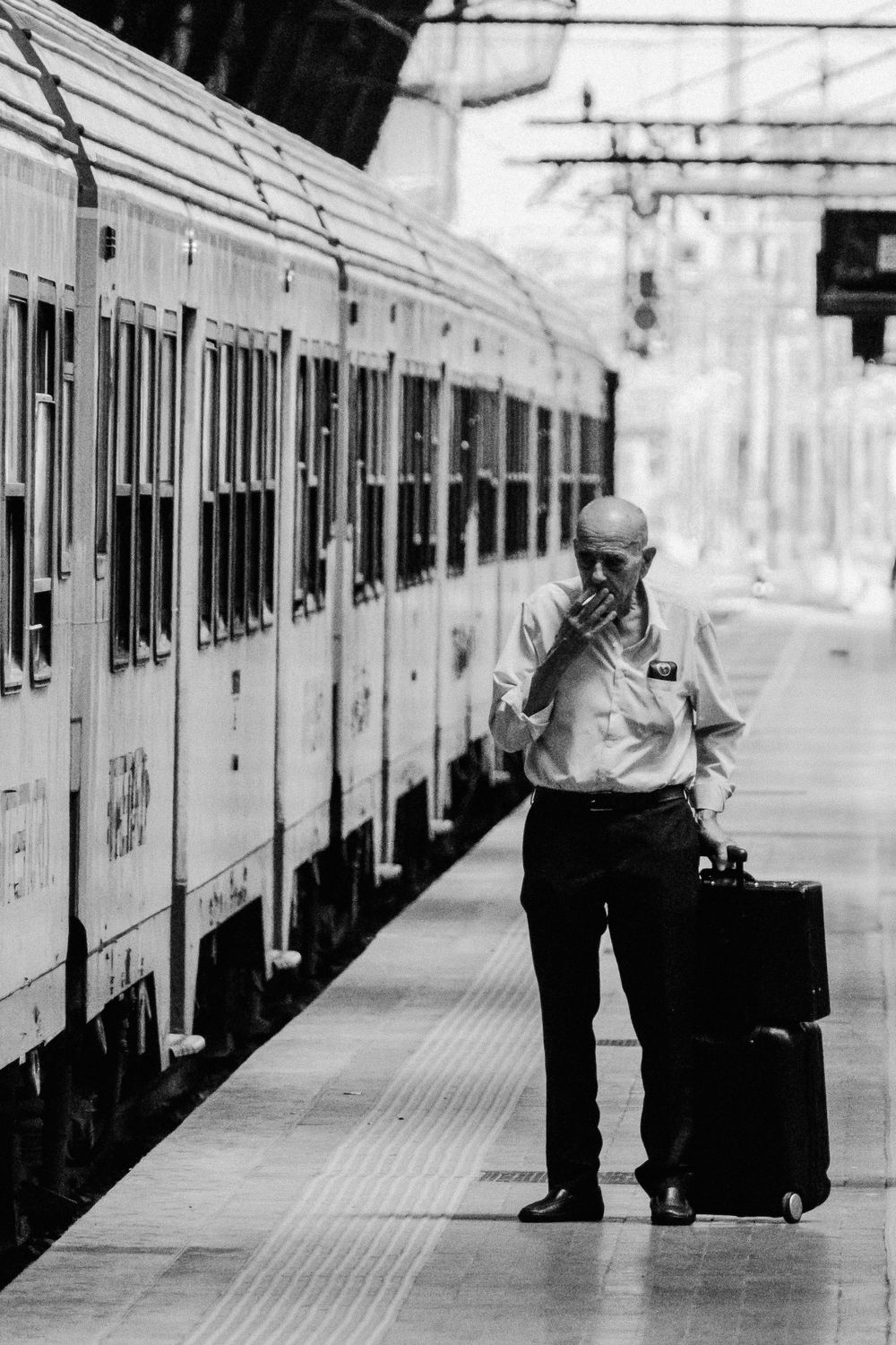 Man with suitcase standing on train platform in black and white urban scene.