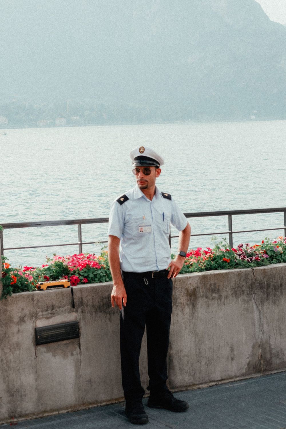 Officer in uniform standing by railing with mountain lake and flowers in background.