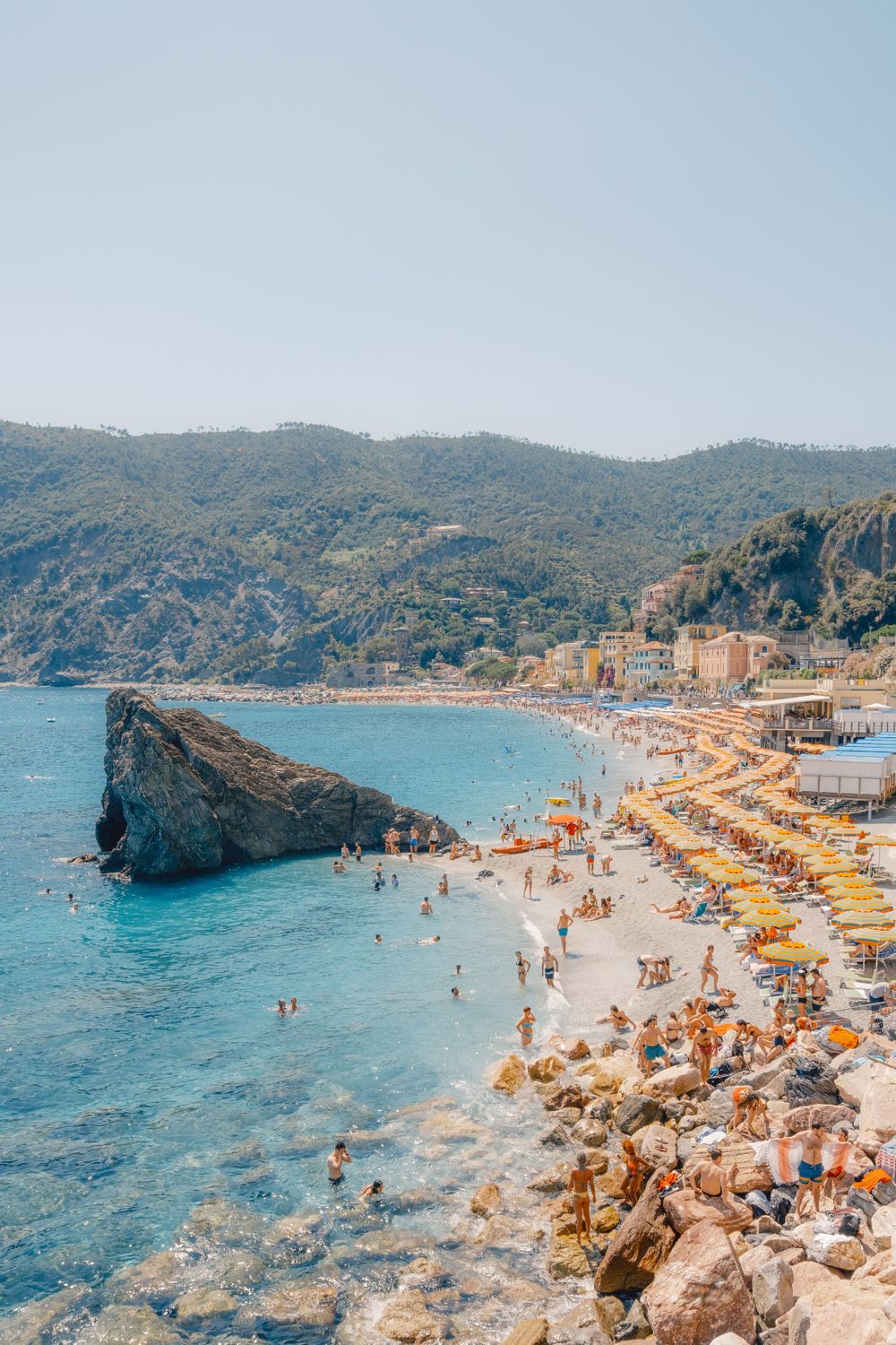 Crowded Mediterranean beach with yellow umbrellas and crystal blue waters along a rocky coastline.