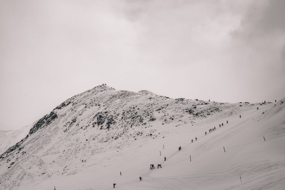 Black and white view of a snow-covered mountain slope with tiny skiers dotting the terrain.