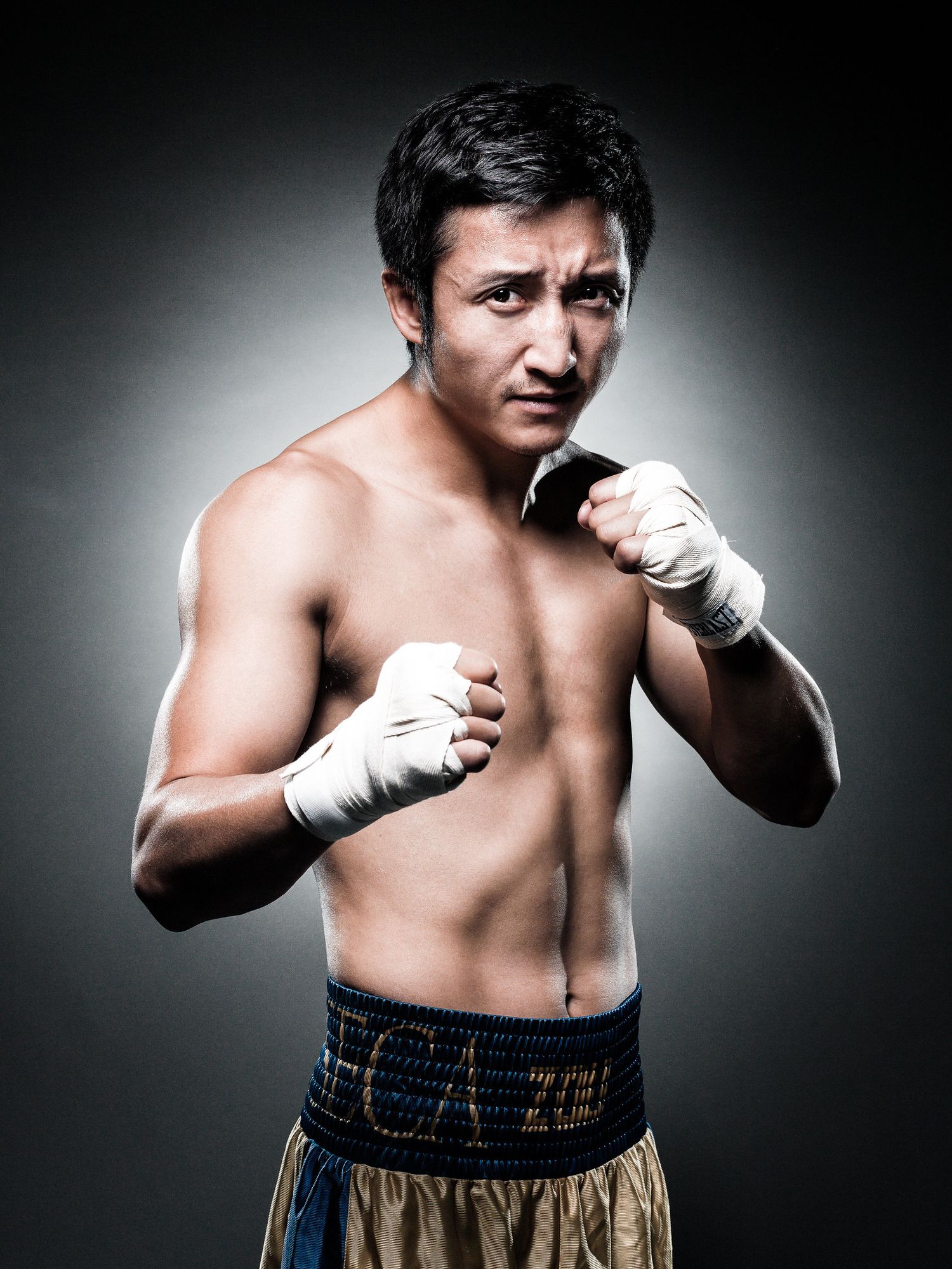 Shirtless boxer in fighting stance with wrapped hands against dark background.