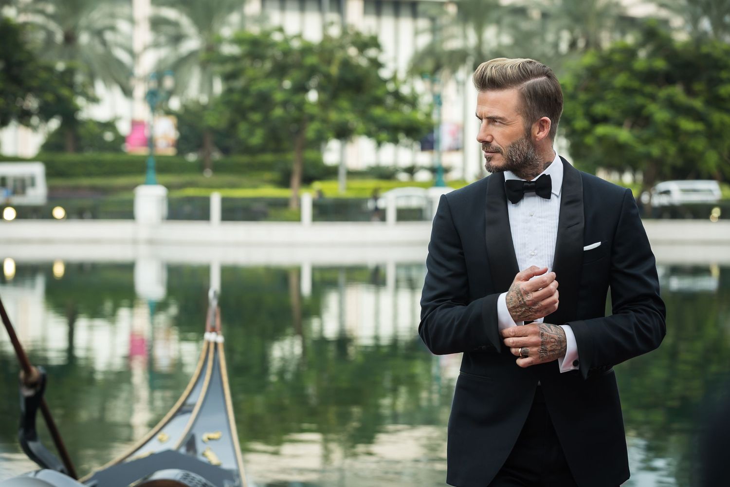 Elegant man in tuxedo adjusts cufflinks by a waterfront setting.