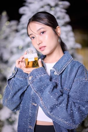 Young Asian woman in a sparkly denim jacket holding a perfume bottle near a snow-covered Christmas tree at night.