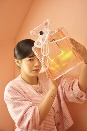 Young Asian woman in pink cardigan examining a large transparent perfume bottle shaped purse with pearl strap.